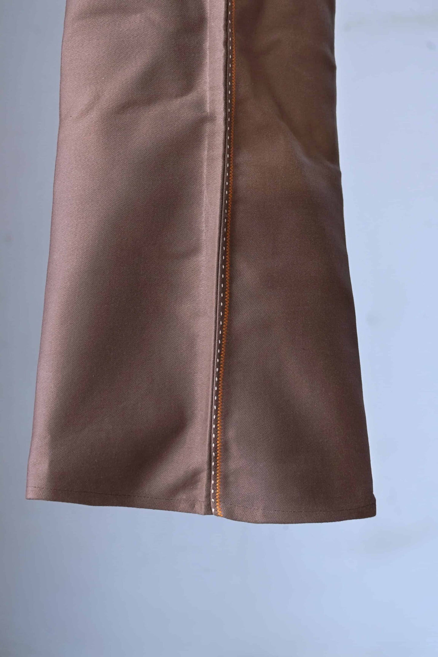 Close-up of the bottom hem of brown pants with orange stitching.
 The pants have a wide leg opening.