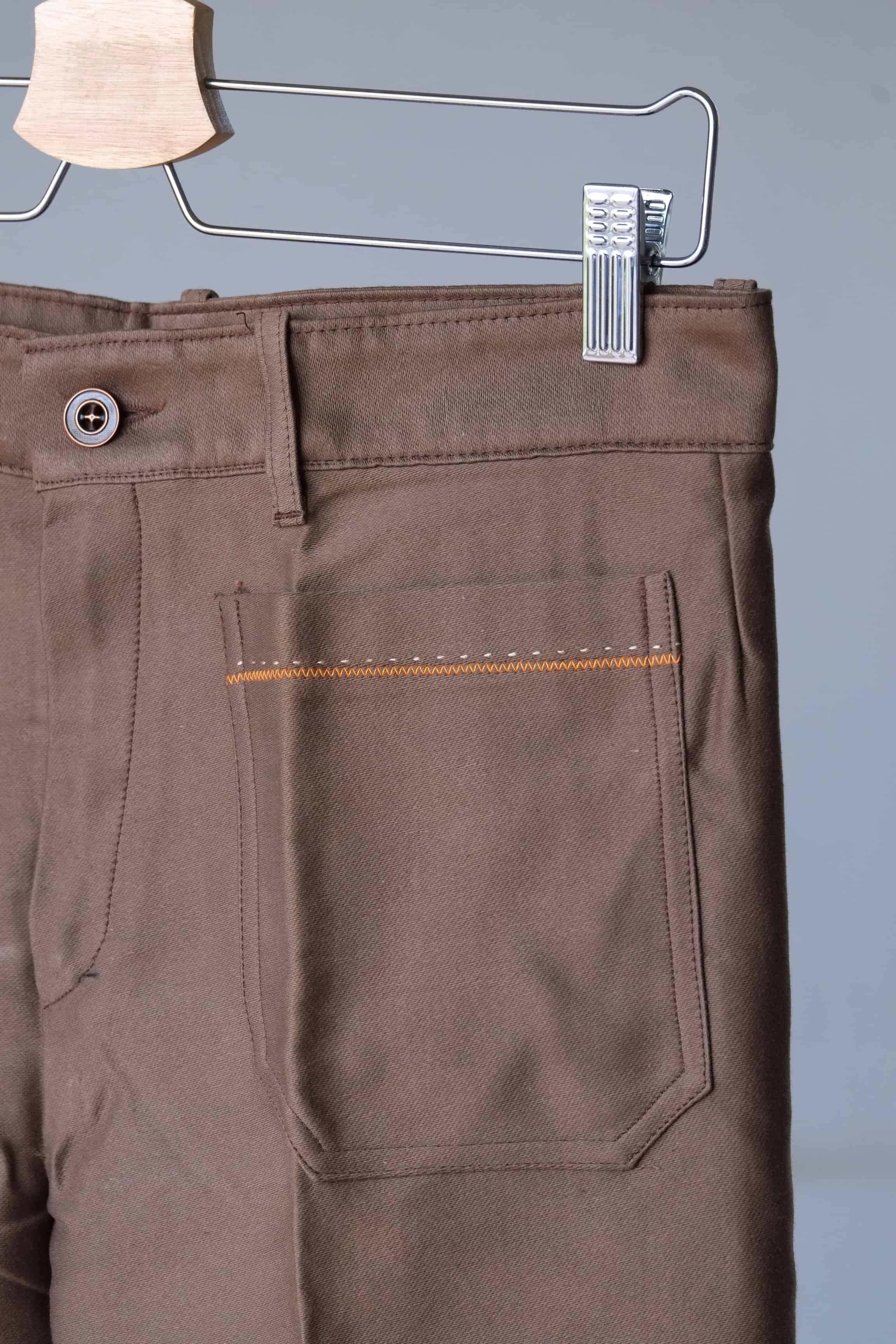 Close-up of the waistband and back pocket of brown pants.
 The pants feature a button closure, belt loops, and orange stitching detailing.