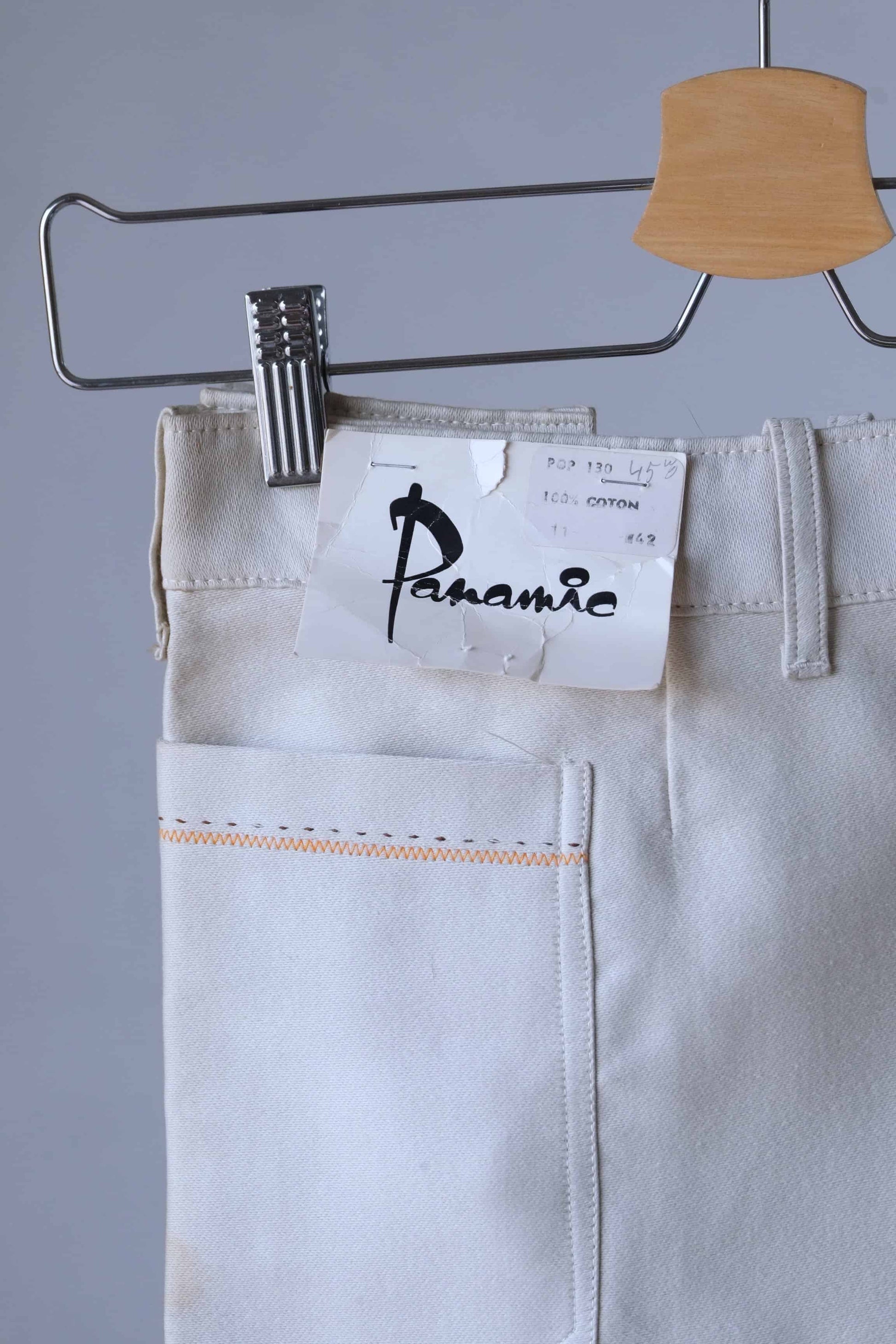 Close-up of a vintage pair of cream-colored pants with a label where the word "Panamic" can be seen. The label indicates they are 100% cotton. The pants feature orange stitching details.