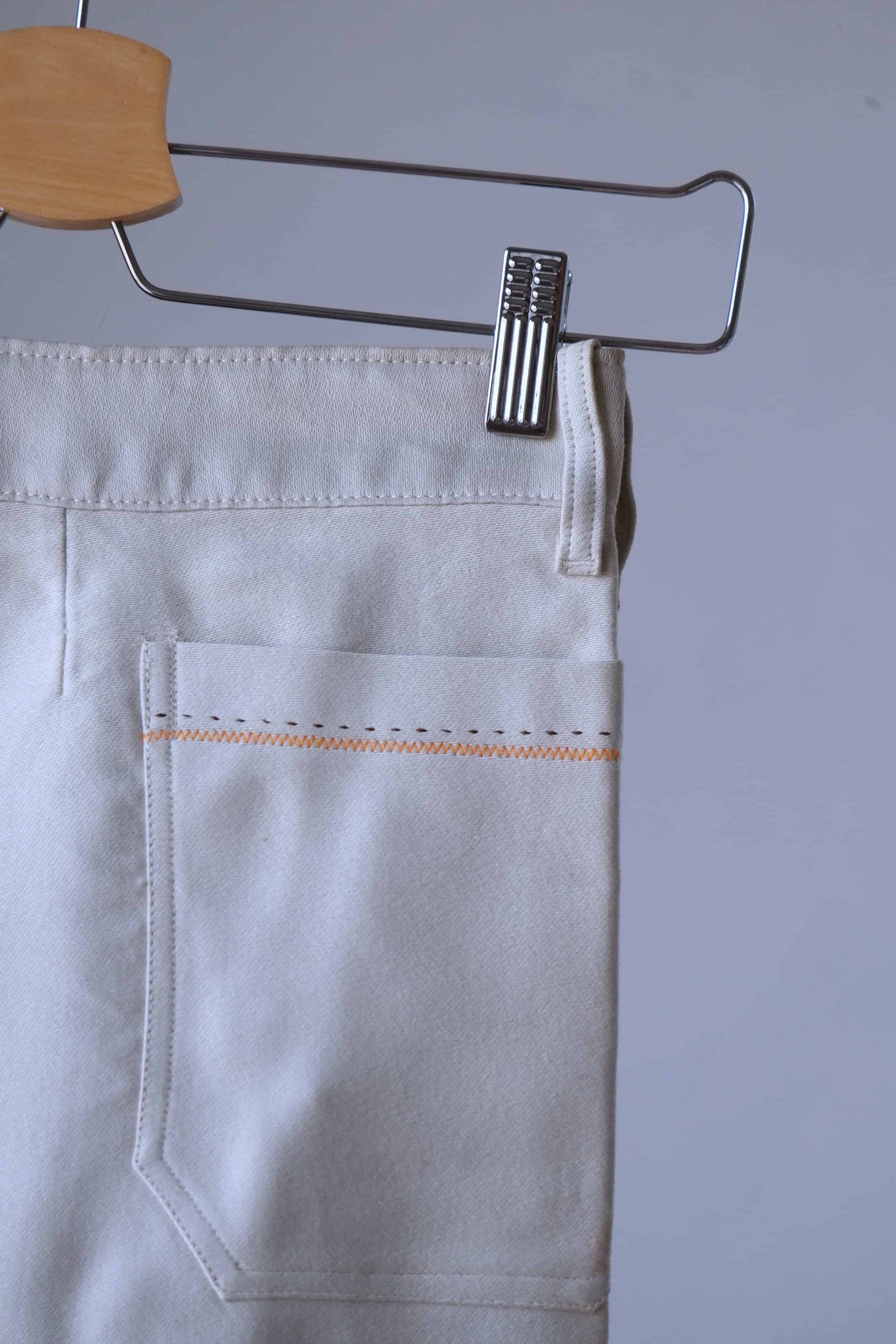 Close-up of the back pocket of cream-colored pants with orange and black stitching. The pants are hanging on a wooden hanger with a small metal clip.