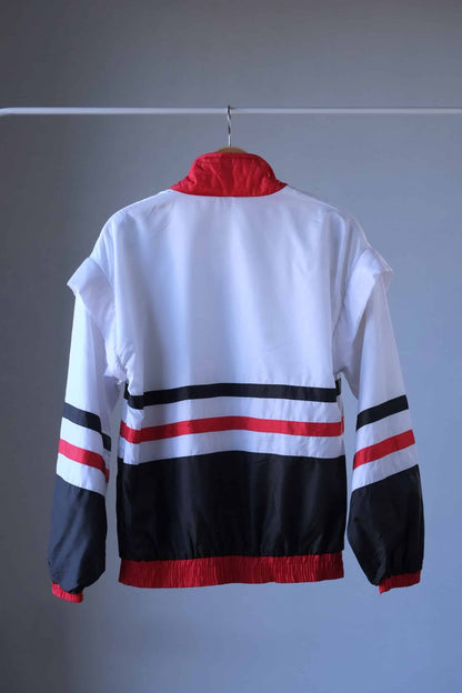 Back view of a white, lightweight Lacoste jacket with red ribbed collar, cuffs, and hem. The jacket has a full zipper and black and red horizontal stripes across the chest. The Lacoste crocodile logo is visible on the left side of the chest. The jacket hangs on a wooden hanger against a neutral gray background.