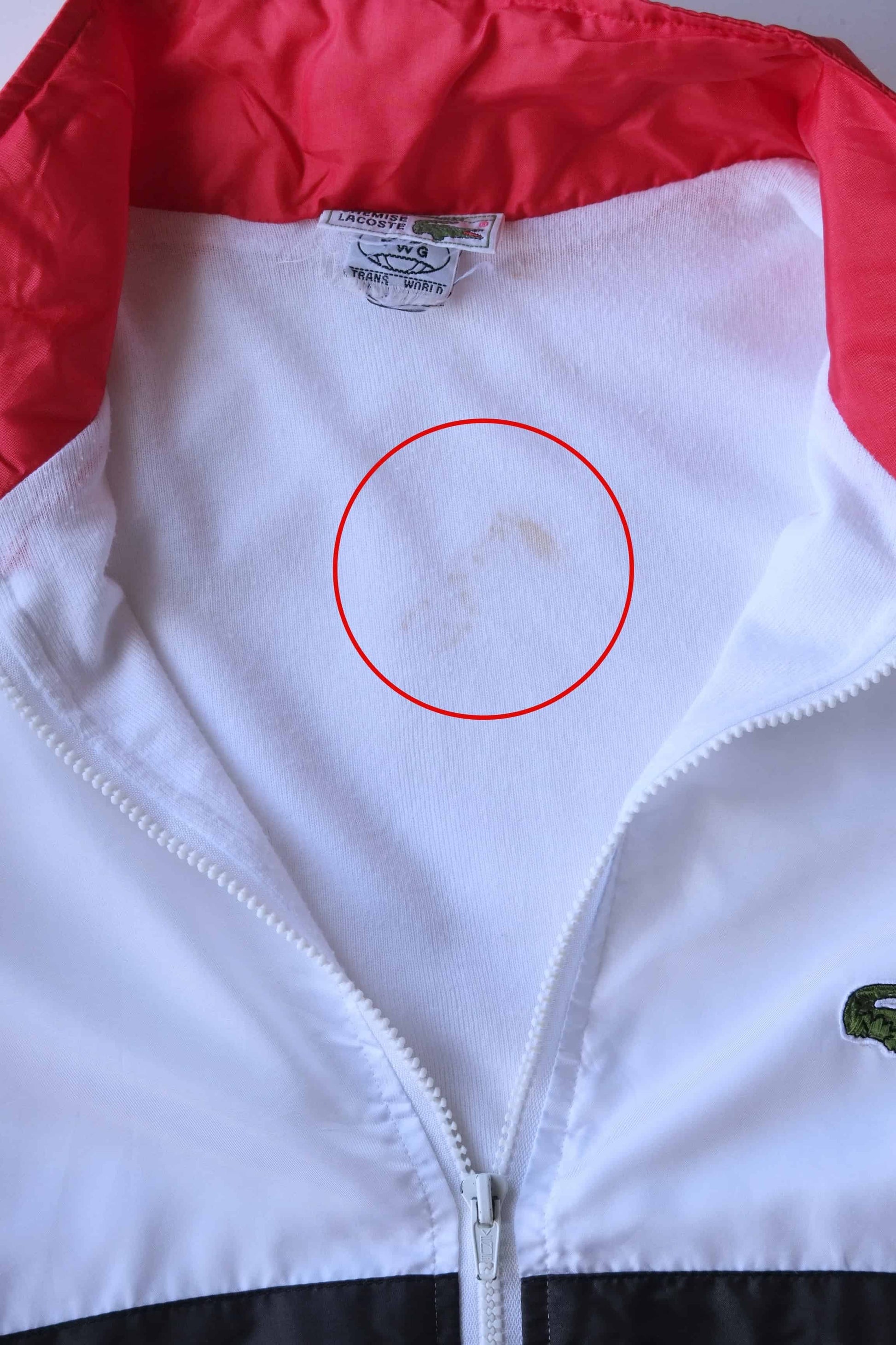 Close-up view of a white Lacoste jacket with a bright red collar, a white zipper, and a tag visible inside the neckline. A red circle is visible and it highlights a light stain.