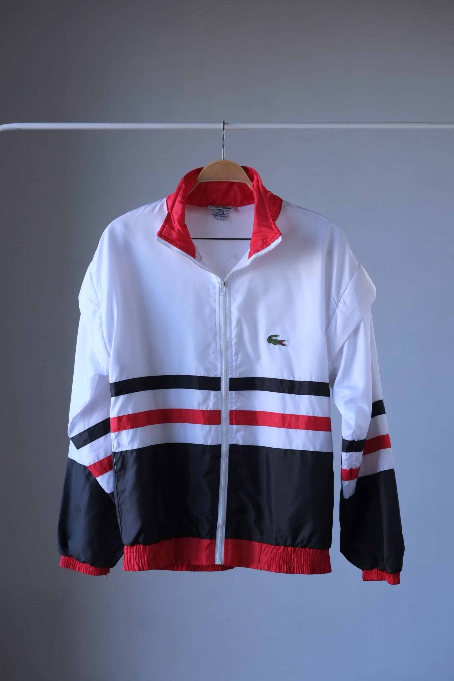 A white, lightweight Lacoste jacket with red ribbed collar, cuffs, and hem. The jacket has a full zipper and black and red horizontal stripes across the chest. The Lacoste crocodile logo is visible on the left side of the chest. The jacket hangs on a wooden hanger against a neutral gray background.