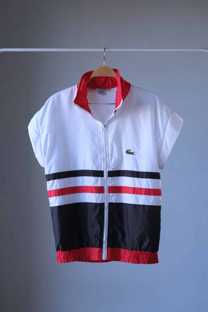 A white, lightweight Lacoste jacket with its sleeves removed. It has a red ribbed collar, cuffs, and hem. The jacket has a full zipper and black and red horizontal stripes across the chest. The Lacoste crocodile logo is visible on the left side of the chest. The jacket hangs on a wooden hanger against a neutral gray background.