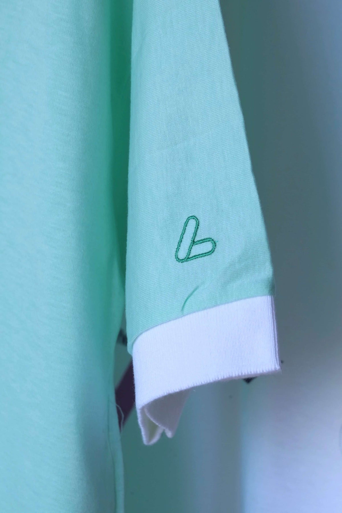 Close-up of white ribbed sleeve cuff and green embroidered Löffler logo on the sleeve of a mint green polo