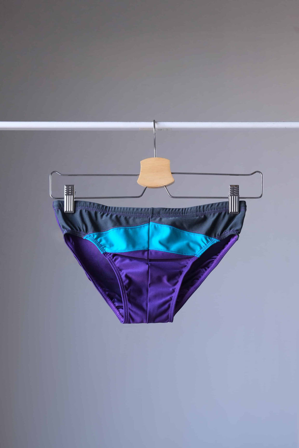 Vintage 1980's men's swim briefs in vibrant purple, teal, and grey color block design, displayed on a hanger.