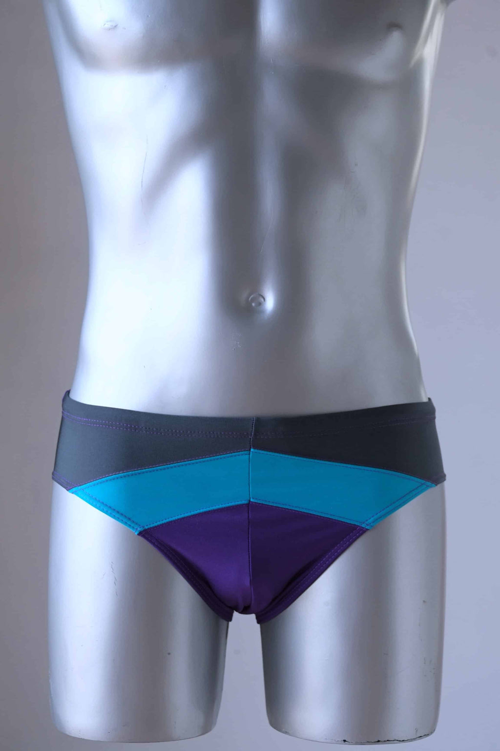 Vintage 1980's men's swim briefs in vibrant purple, teal, and grey color block design, worn by a silver mannequin.