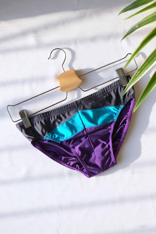 Vintage 1980's retro color-block swimming briefs in vibrant purple, teal, and grey, displayed on a hanger against a white background.