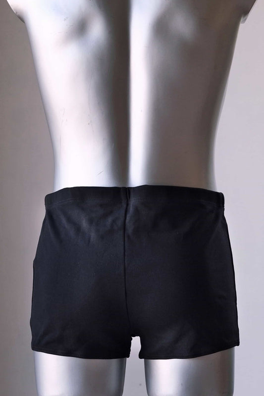 Back view of a vintage 1960s men's square-cut swim trunks on a silver mannequin. The trunks feature subtle vertical stitched panel lines and a small gold metal logo badge on the left hip.