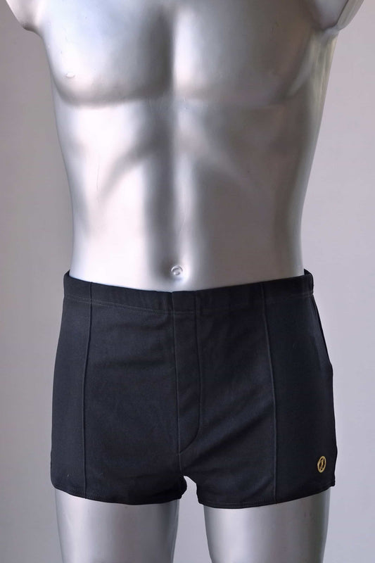 Front view of a vintage 1960s men's square-cut swim trunks on a silver mannequin. The trunks feature subtle vertical stitched panel lines and a small gold metal logo badge on the left hip.