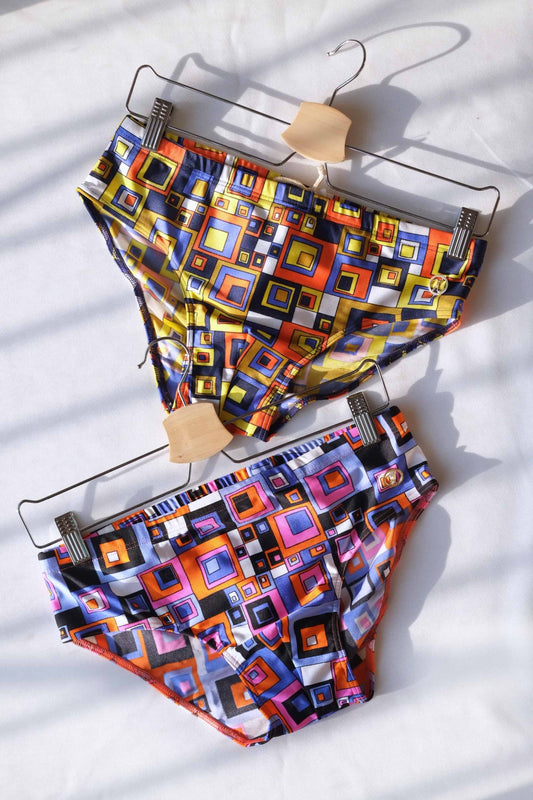 Two pairs of vintage 80s retro geometric square swim briefs with vibrant, colorful patterns hanging on hangers against a white background with soft shadows.