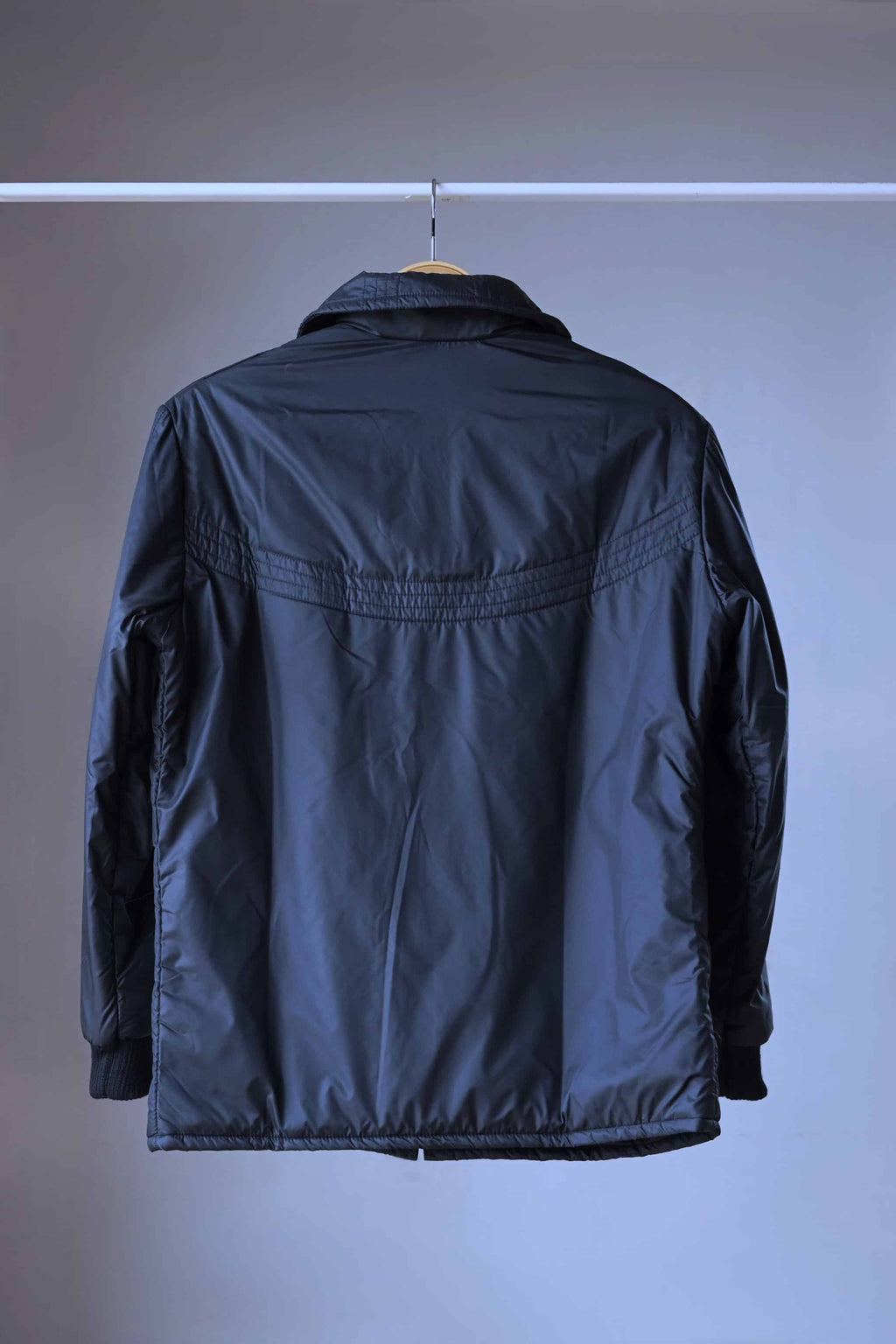 Back view of the vintage 70s Mossant Rangoon black ski jacket hanging on a wooden hanger, featuring a horizontal quilted yoke design and ribbed cuffs.