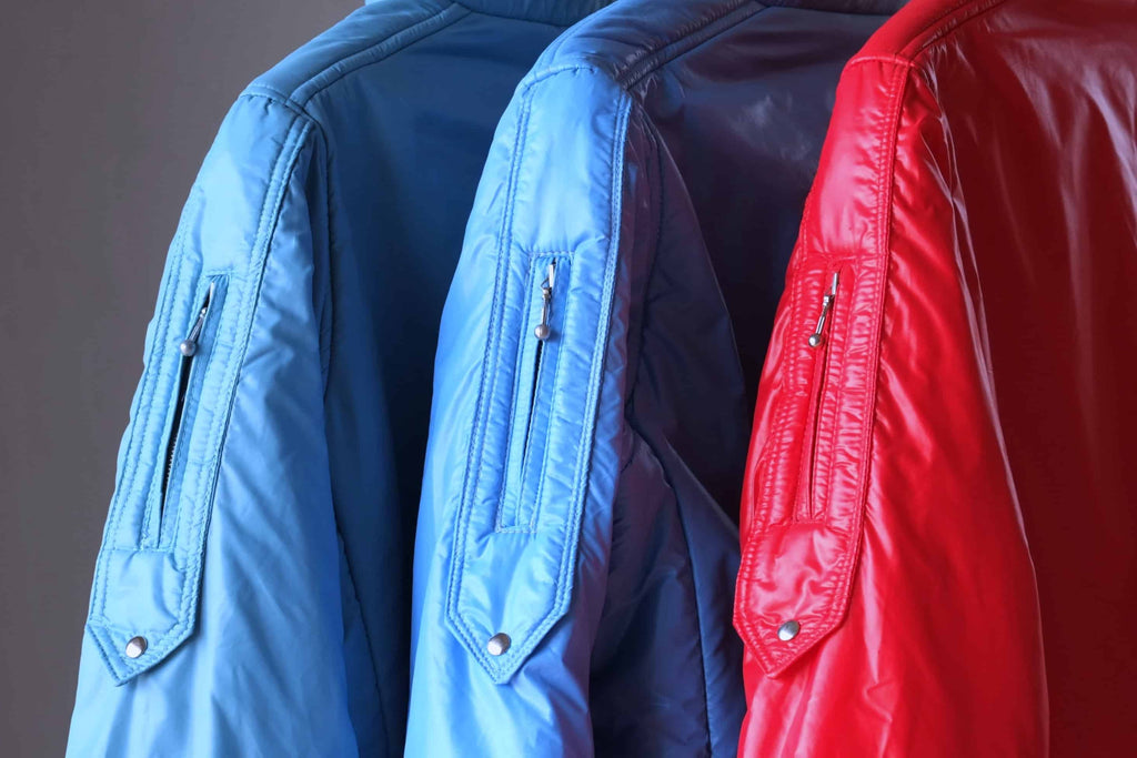 A side-profile close-up of three vintage Mossant ski jackets in sky blue, steel blue, and red. The focus is on the upper arm utility pockets, featuring vertical zippers with silver-tone ball pull tabs and a pointed flap base secured with a metal snap.