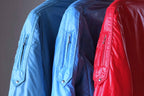 A side-profile close-up of three vintage Mossant ski jackets in sky blue, steel blue, and red. The focus is on the upper arm utility pockets, featuring vertical zippers with silver-tone ball pull tabs and a pointed flap base secured with a metal snap.