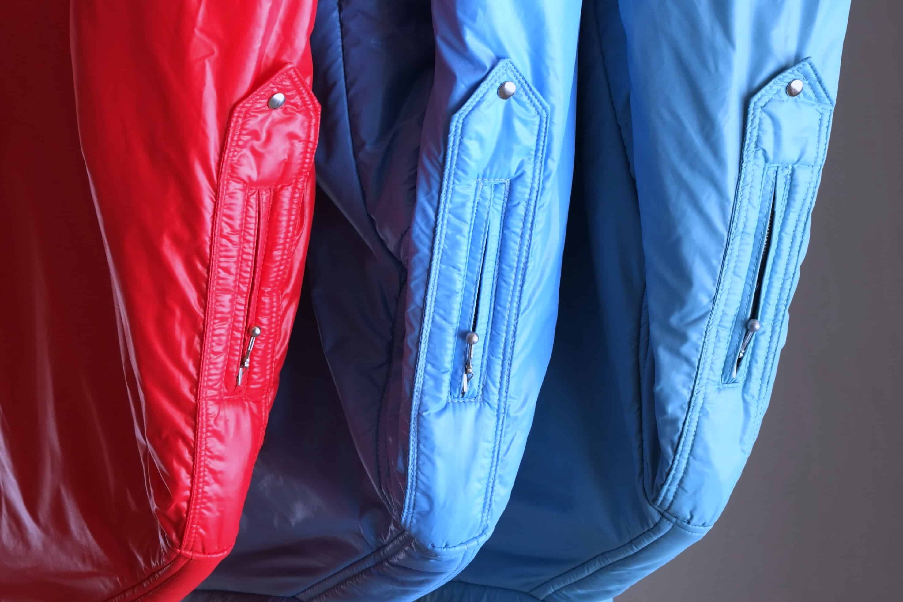 A side-profile close-up of three vintage Mossant ski jackets in sky blue, steel blue, and red. The focus is on the upper arm utility pockets, featuring vertical zippers with silver-tone ball pull tabs and a pointed flap base secured with a metal snap.