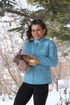 A woman outdoors in a snowy, wooded setting wearing the sky blue Mossant vintage ski jacket. The jacket shows a slim, fitted silhouette, a high snap-button collar, and multiple technical zippered pockets.