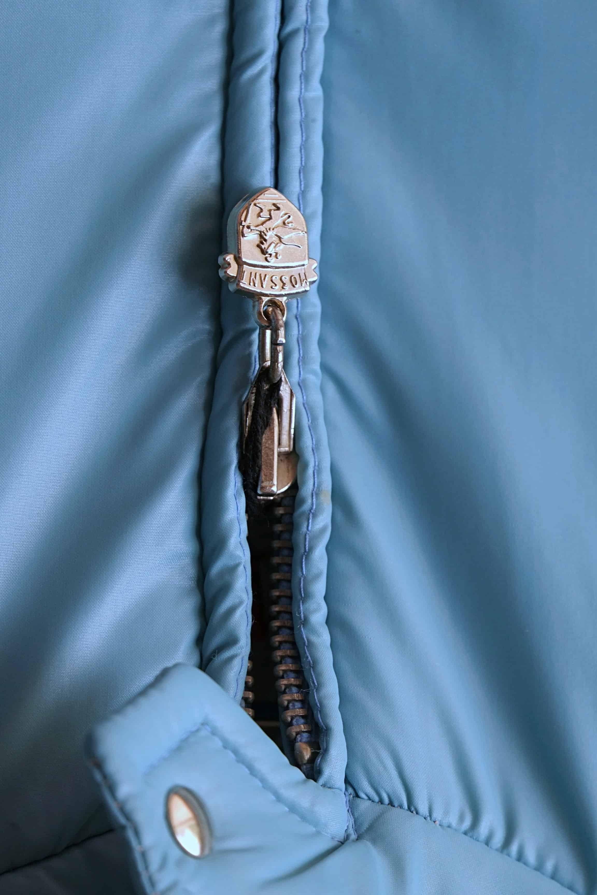 A tight macro shot of the steel blue Mossant ski jacket’s central zipper. The silver-tone zipper pull is embossed with the Mossant brand crest, featuring a winged horse (Pegasus) design.