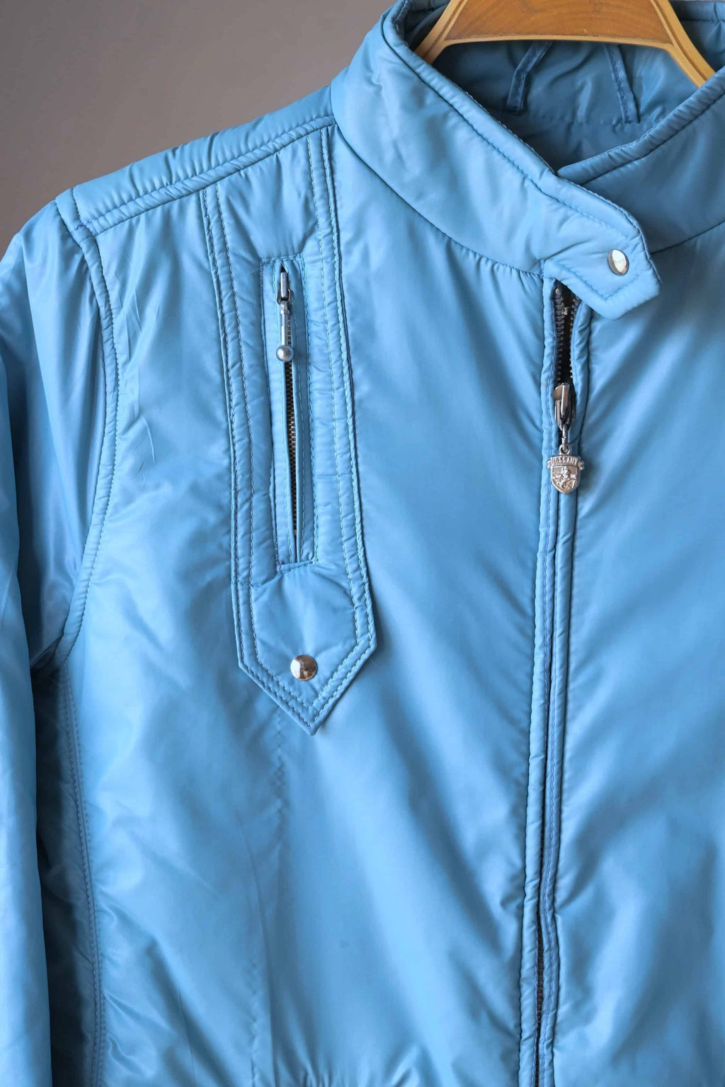 Detailed shot of the sky blue Mossant jacket's neckline and zipper. The high collar is secured with a silver snap, and the zipper pull features the signature Mossant Pegasus crest.