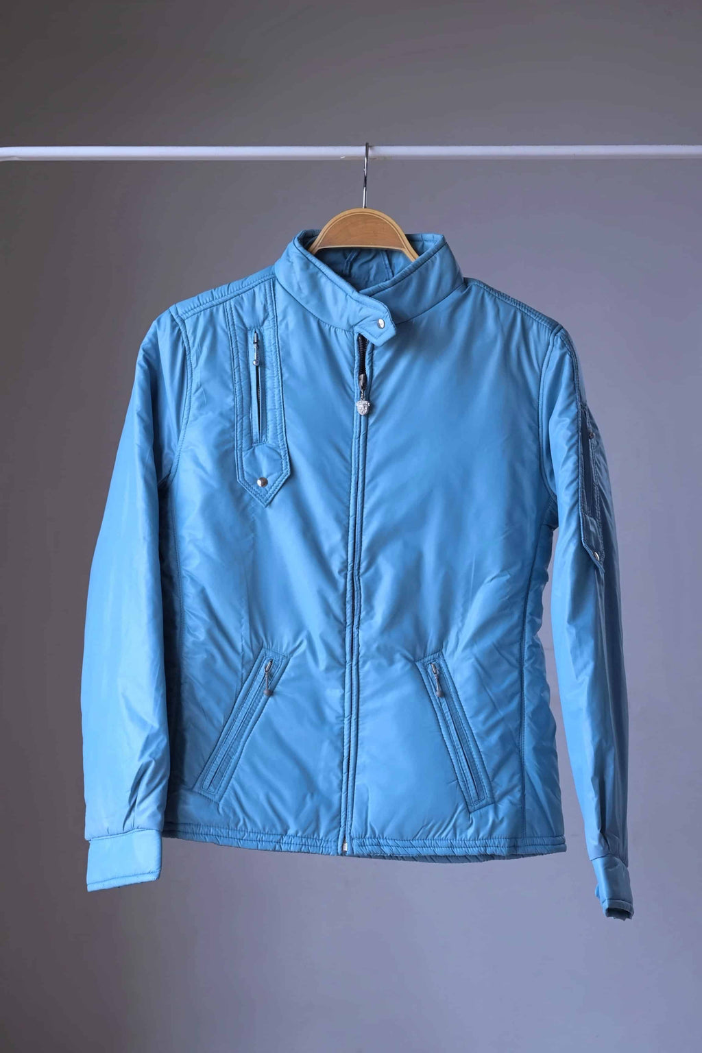 Full front view of the steel blue Mossant vintage 70s ski jacket on a wooden hanger. The jacket features a slim, athletic fit, technical zippered pockets on the chest and waist, and a snap-closure high collar.
