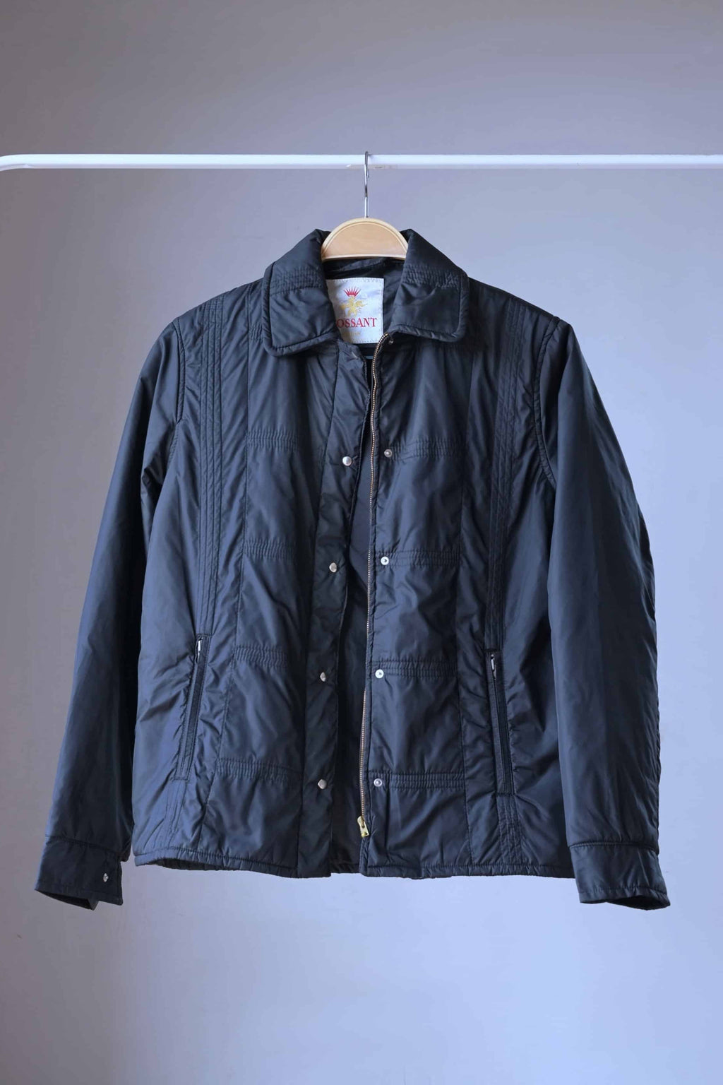 Front view of a vintage 1970s Mossant black ski jacket on a wooden hanger, showing a grid-quilted pattern, shirt-style collar, and a silver snap-button storm flap over a zipper.