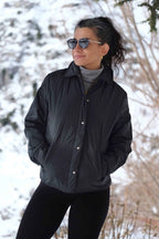 A woman outdoors in a snowy landscape wearing the black 70s Mossant jacket, styled with sunglasses and a grey turtleneck.
