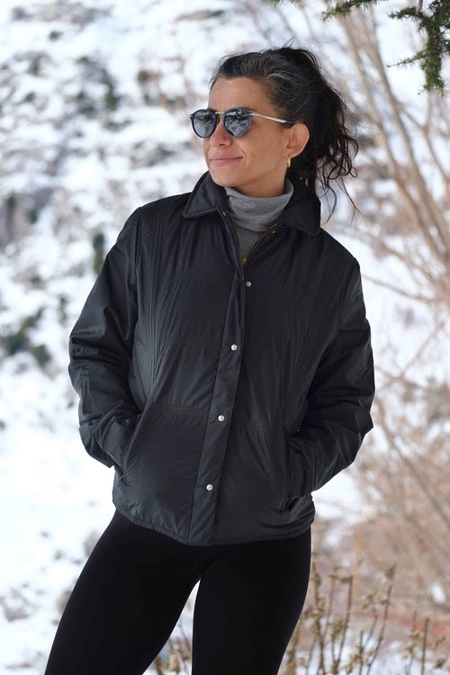 A woman outdoors in a snowy landscape wearing the black 70s Mossant jacket, styled with sunglasses and a grey turtleneck.