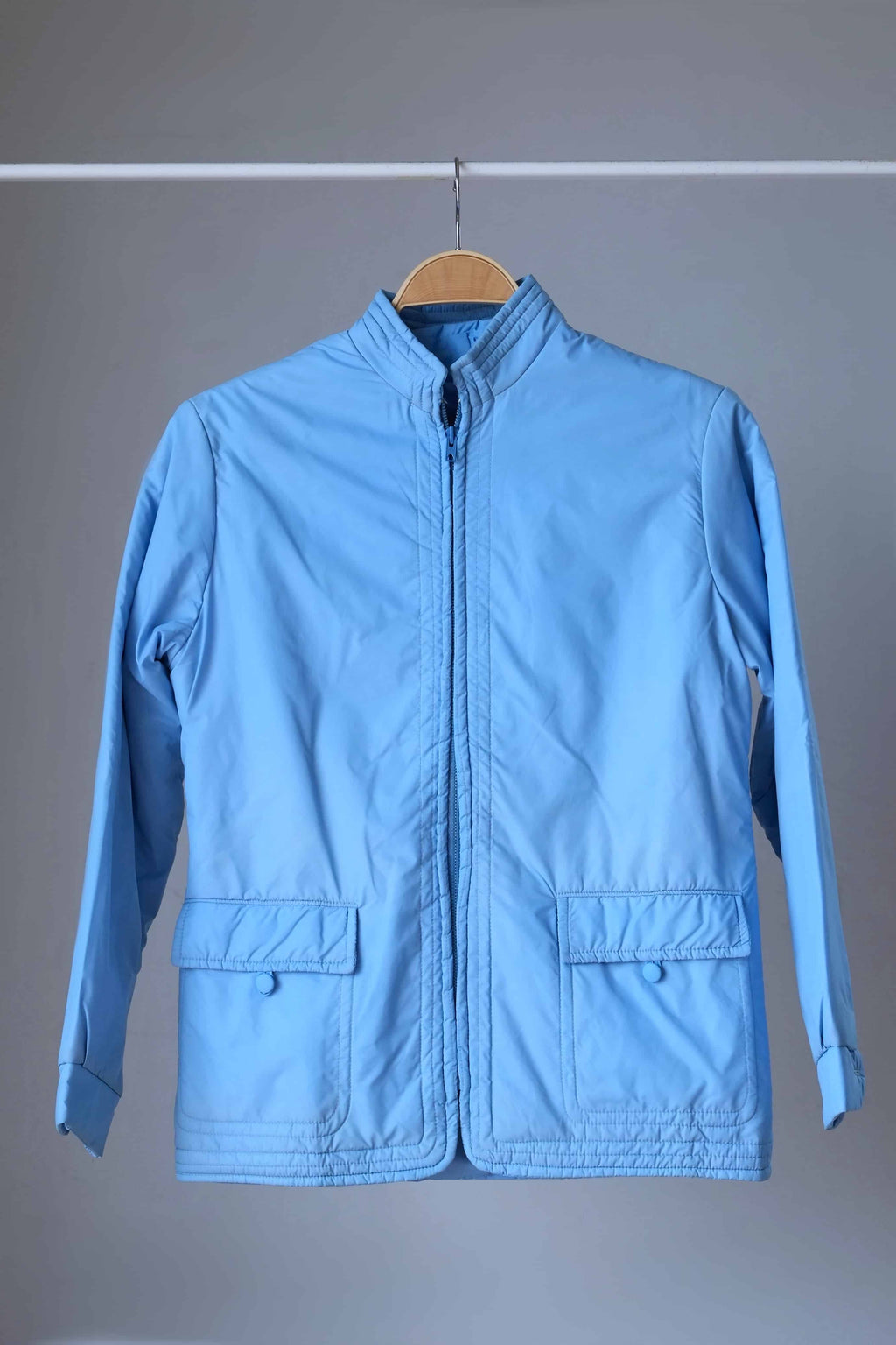 A front-facing shot of an azure blue 1970s Mossant ski jacket on a wooden hanger, featuring a full-length zipper and two large front flap pockets.