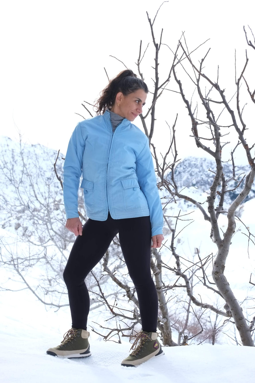 A model posing in the snow wearing the azure blue Mossant ski jacket, styled with black leggings and hiking boots to show the jacket's hip-length fit.