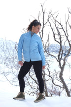 A model posing in the snow wearing the azure blue Mossant ski jacket, styled with black leggings and hiking boots to show the jacket's hip-length fit.