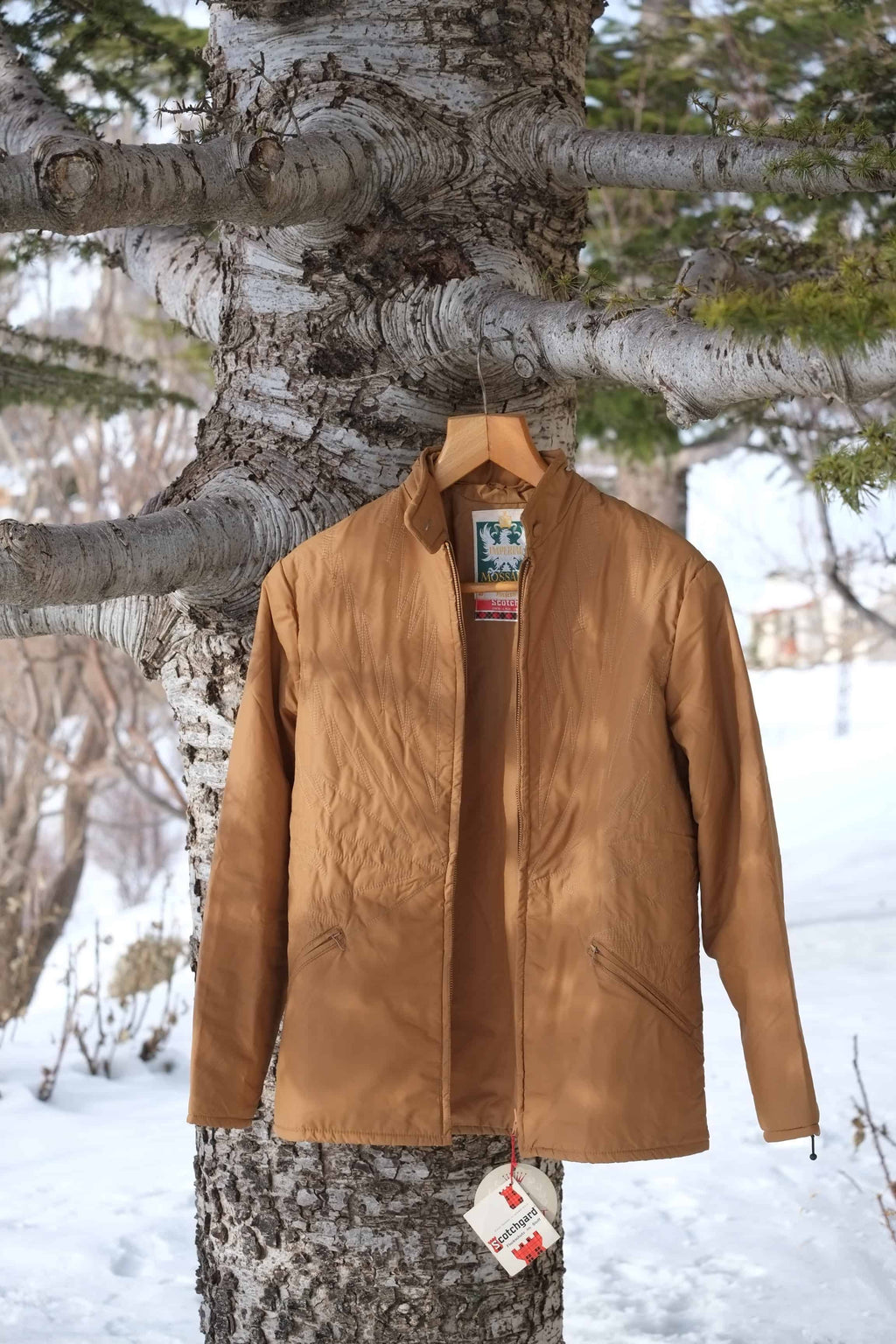 A vintage MOSSANT 70s camel brown quilted ski jacket hanging on a wooden hanger against a snowy outdoor background.