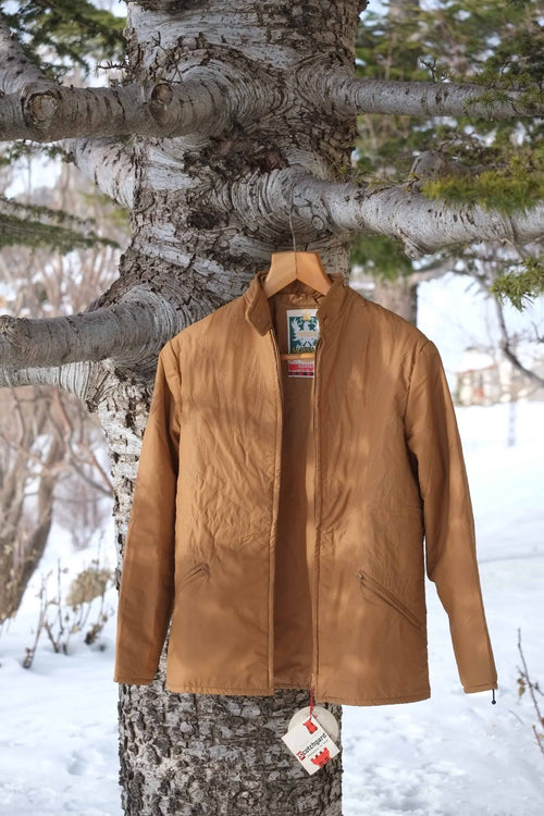 A vintage MOSSANT 70s camel brown quilted ski jacket hanging on a wooden hanger against a snowy outdoor background.