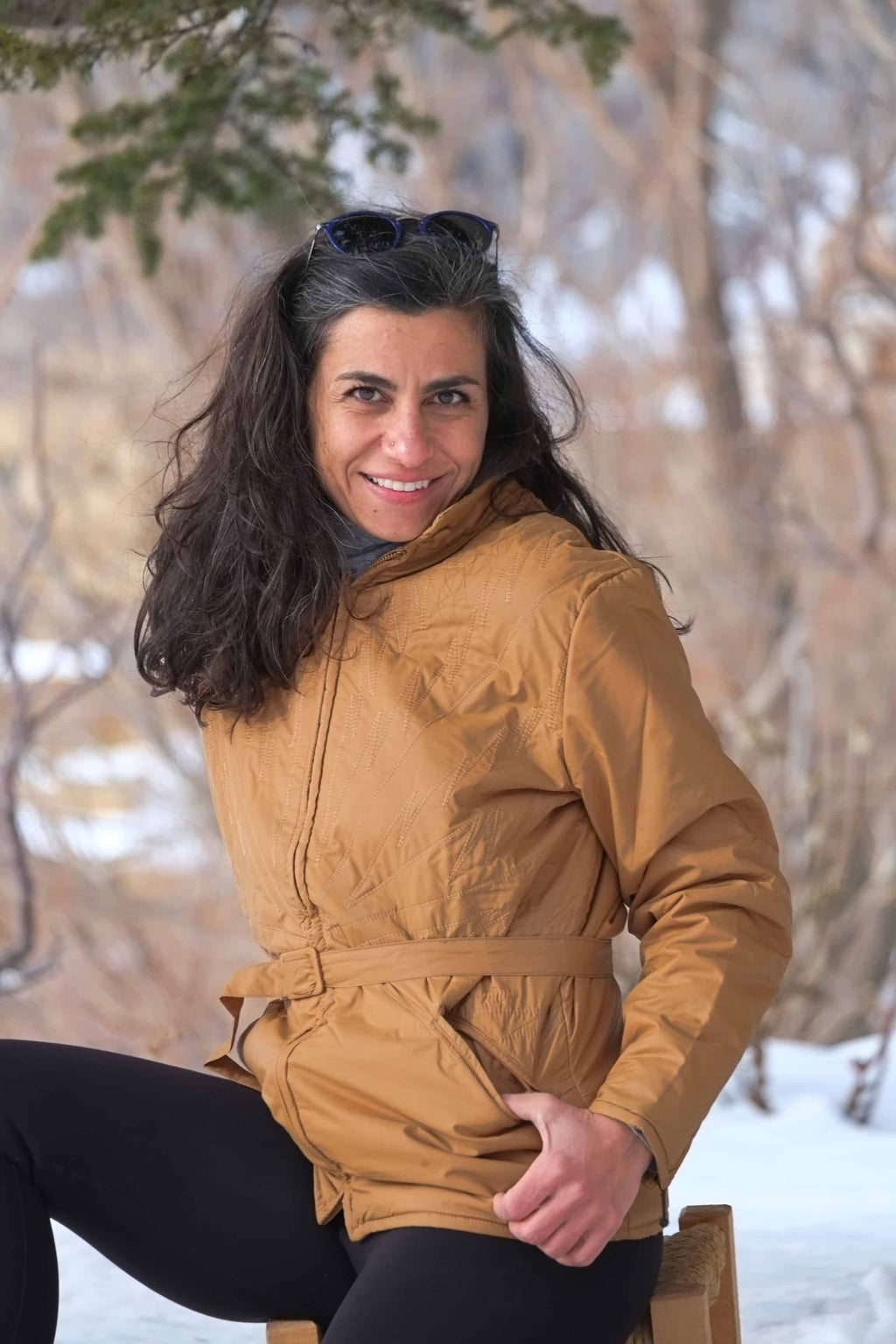 A woman smiling while wearing the belted Mossant camel brown ski jacket in a snowy outdoor setting.