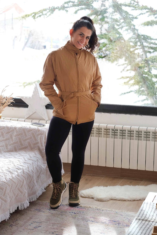Full-length view of a woman wearing the camel brown Mossant ski jacket with a waist-cinching belt and winter boots.