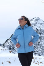 A front-facing shot of the woman in the snow wearing the zipped-up sky-blue Mossant jacket and sunglasses, demonstrating the jacket's vibrant color and athletic silhouette.