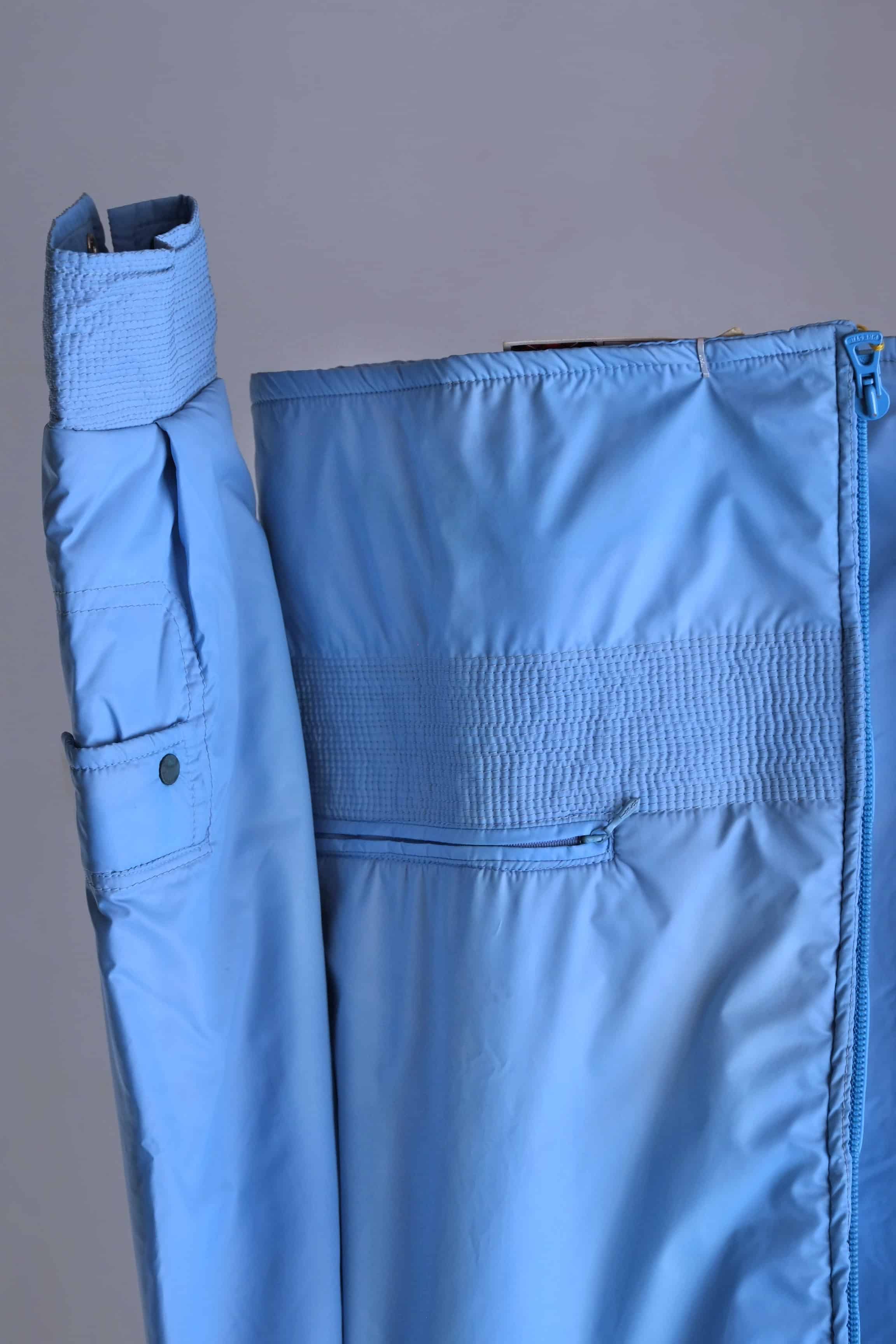 A close-up of the jacket's lower front and sleeve, showing a zippered hip pocket, smocked waist band, and a small utility pocket on the forearm with a snap closure.