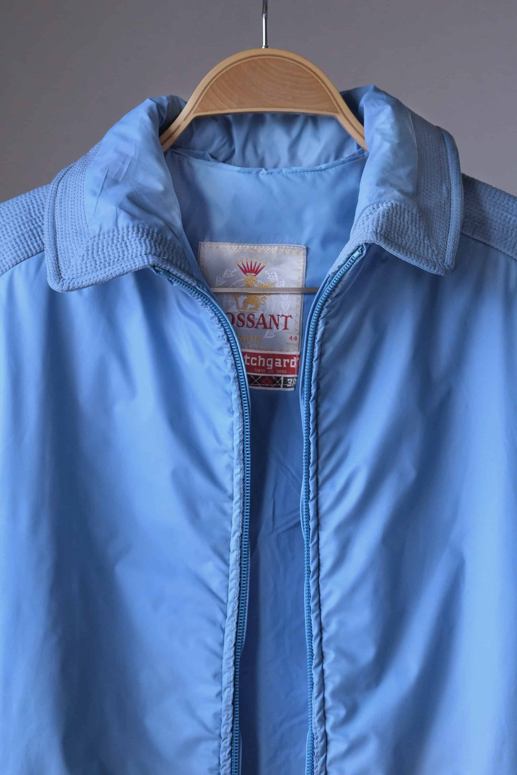 A close-up of the open neckline of the sky-blue jacket, clearly showing the "Mossant Paris" brand label and an additional "Scotchgard" fabric care tag.