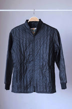 A black Mossant vintage 70s vertical quilted ski jacket hanging on a wooden hanger against a neutral grey background. The jacket has a full-length front zipper, two zippered hand pockets, and a stand-up collar.