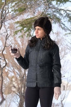 A person outdoors in the snow wearing a black 1970s Mossant quilted ski jacket, styled with a dark faux fur hat and pink knit gloves. The vertical stitching and lightweight puffer design are clearly visible.