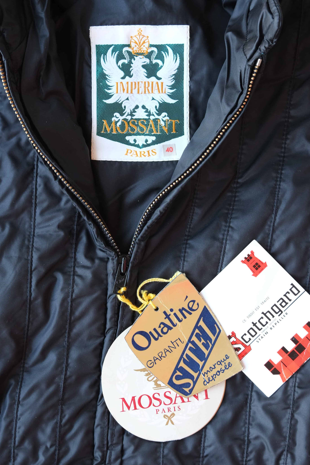 Close-up of the interior neck labels and original paper hangtags for a Mossant Paris jacket. The labels include a green and white "Imperial Mossant Paris" woven tag (size 40), a "Scotchgard" stain repeller tag, and a "Sitel Ouatiné" brand tag.