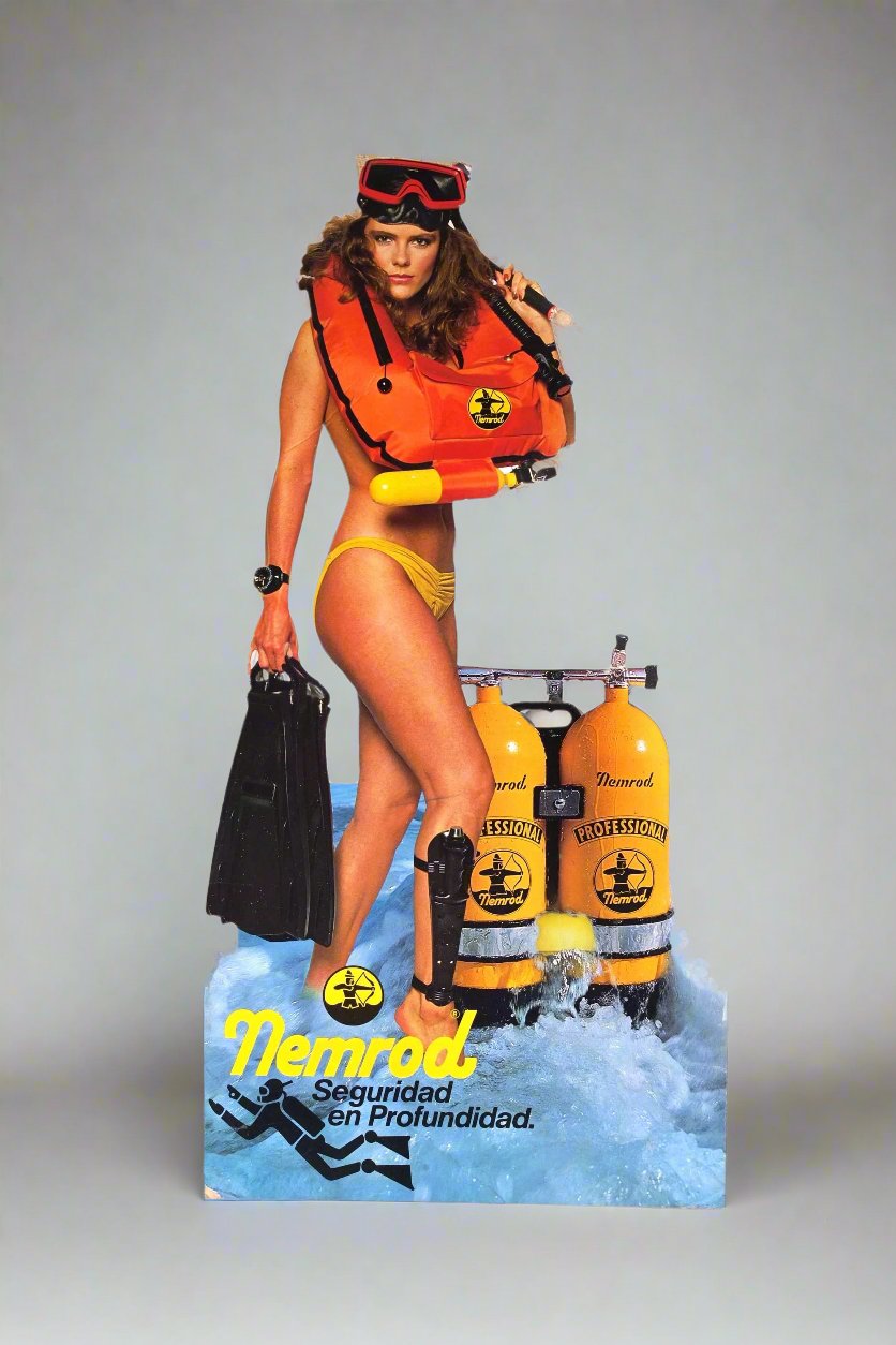 Front view of a Vintage Nemrod scuba diving advertising standee featuring a female model in yellow swimwear, holding black diving fins and wearing an orange buoyancy vest with the Nemrod logo. She stands beside two yellow 'Professional Nemrod' scuba tanks on a blue wave-themed base with the text 'Nemrod Seguridad en Profundidad' in bold yellow and black letters.