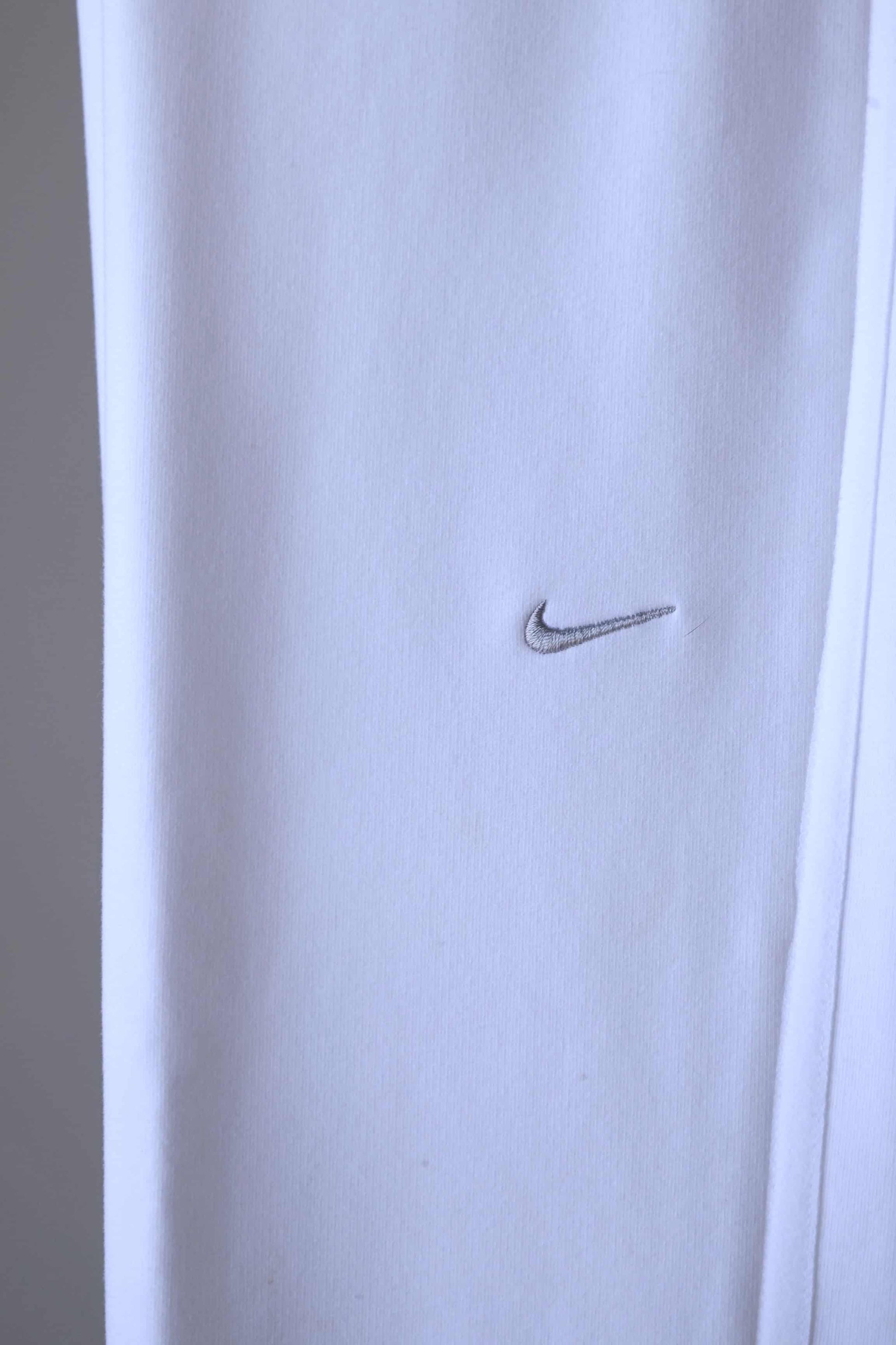 Close-up of a silver Nike swoosh logo embroidered on the right hip of white pants.