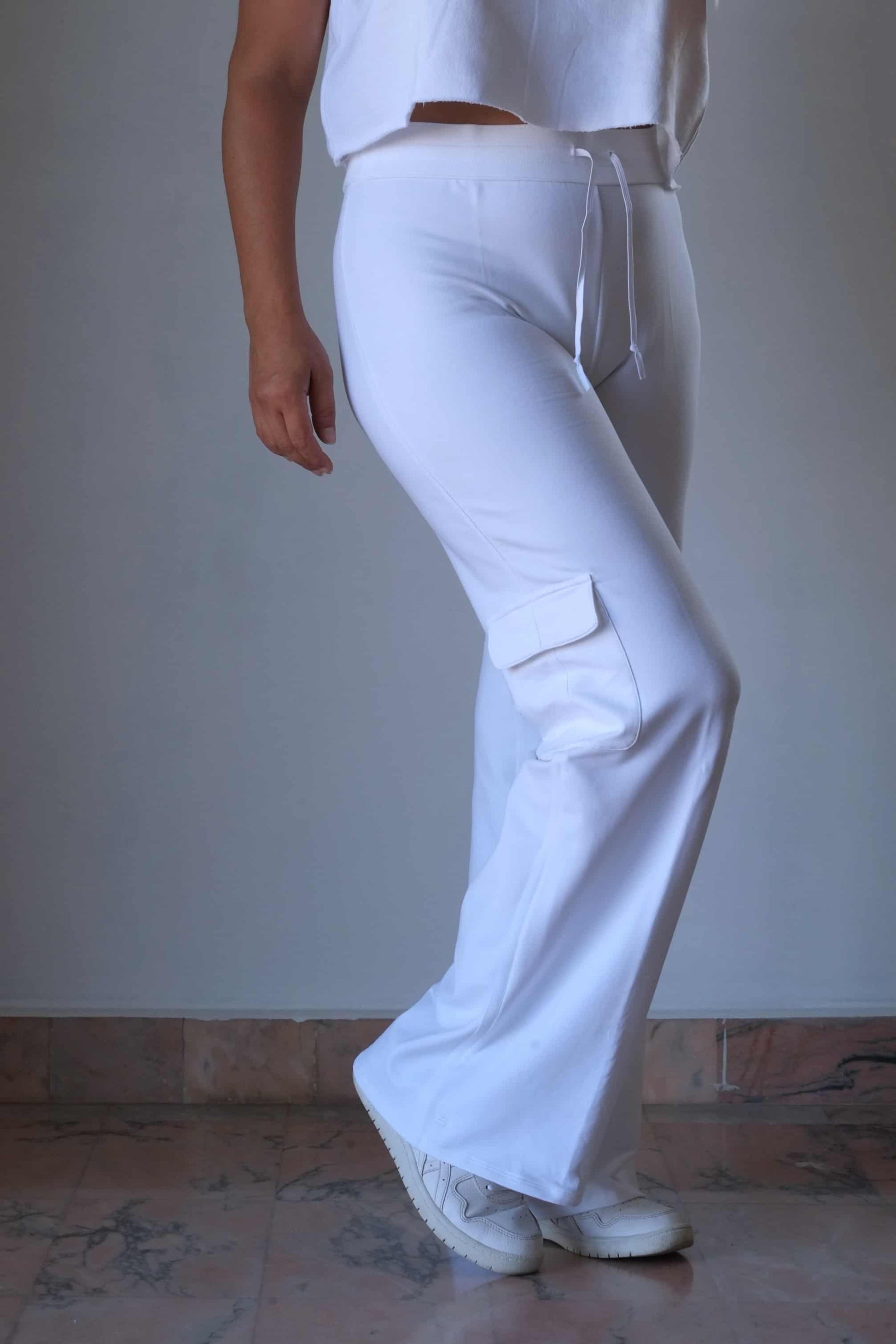 A model flexing their right leg, wearing white Nike flared pants from the 2000s era, featuring a drawstring waist and subtle cargo pockets, standing indoors on a marble floor.