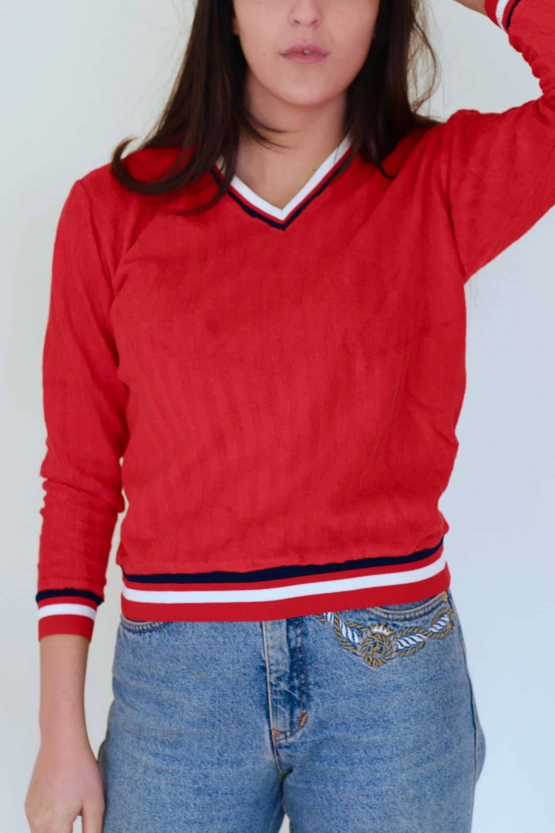 Close-up of a woman wearing a red vintage 70s V-neck sweater with retro striped neckline and cuffs, paired with high-waisted embroidered blue jeans, against a plain white background.