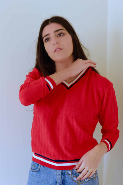 Model wearing a red vintage 70s V-neck sweater with striped ribbed neckline and cuffs, paired with high-waisted blue jeans featuring embroidered details, standing against a plain white background.