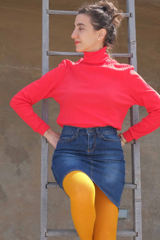 Woman wearing a vibrant red ribbed turtleneck top paired with a denim skirt and mustard-yellow tights, standing confidently against a wooden ladder with a neutral-toned background.