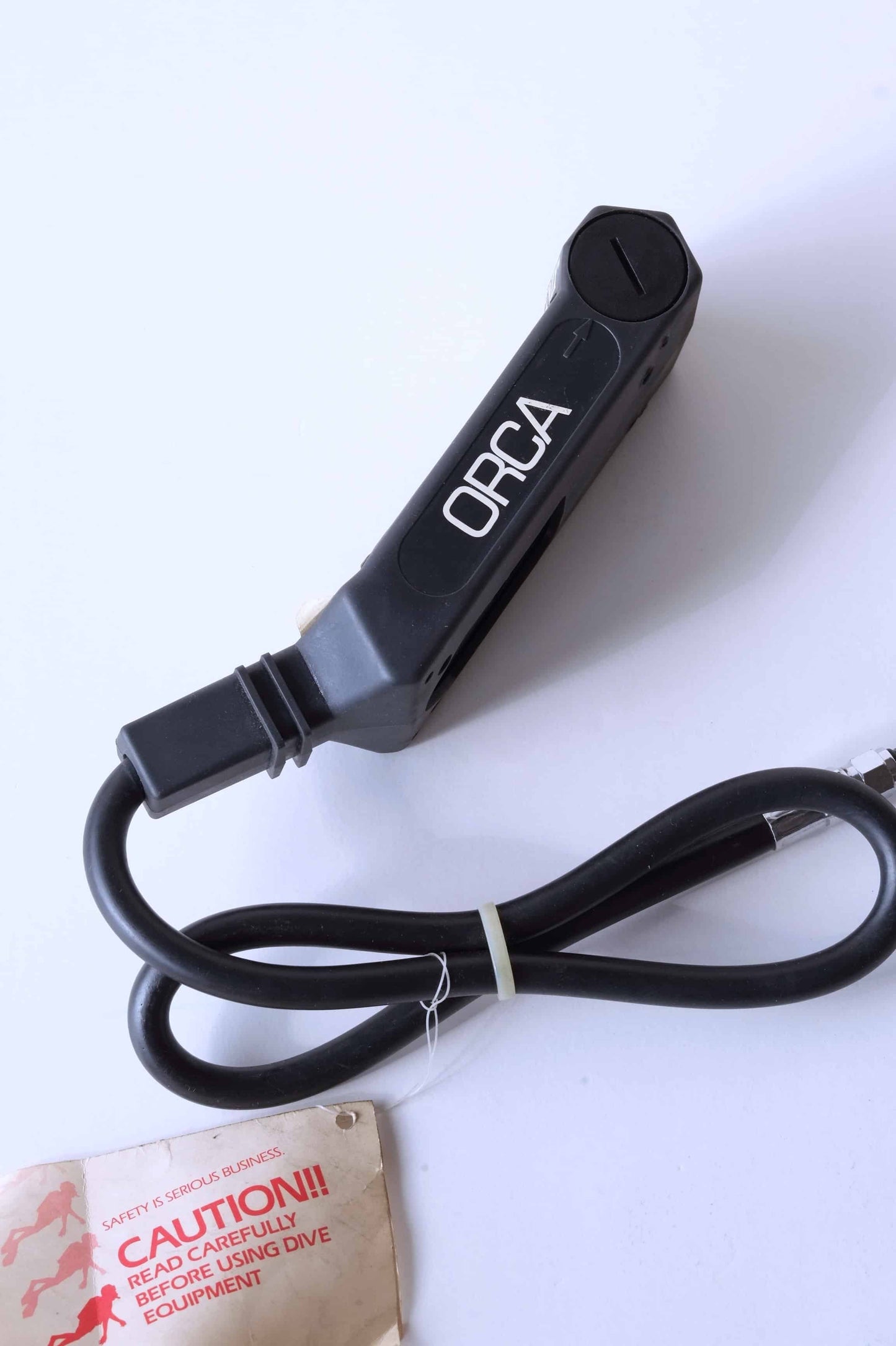 Right side view of a ORCA PHOENIX Air Integrated Scuba Dive Computer displayed on a neutral background. The tube that is attached to the device is tied and a label is hanging from it.