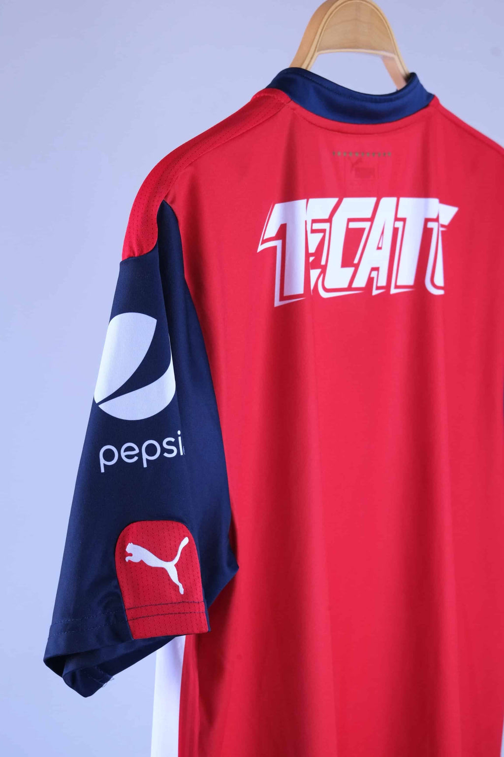 Close-up view of the back shoulder and navy blue sleeve, detailing the white "TECATE" graphic text across the upper back, a white "pepsi" logo printed on the side of the sleeve, and a red patch with a white Puma logo near the cuff.