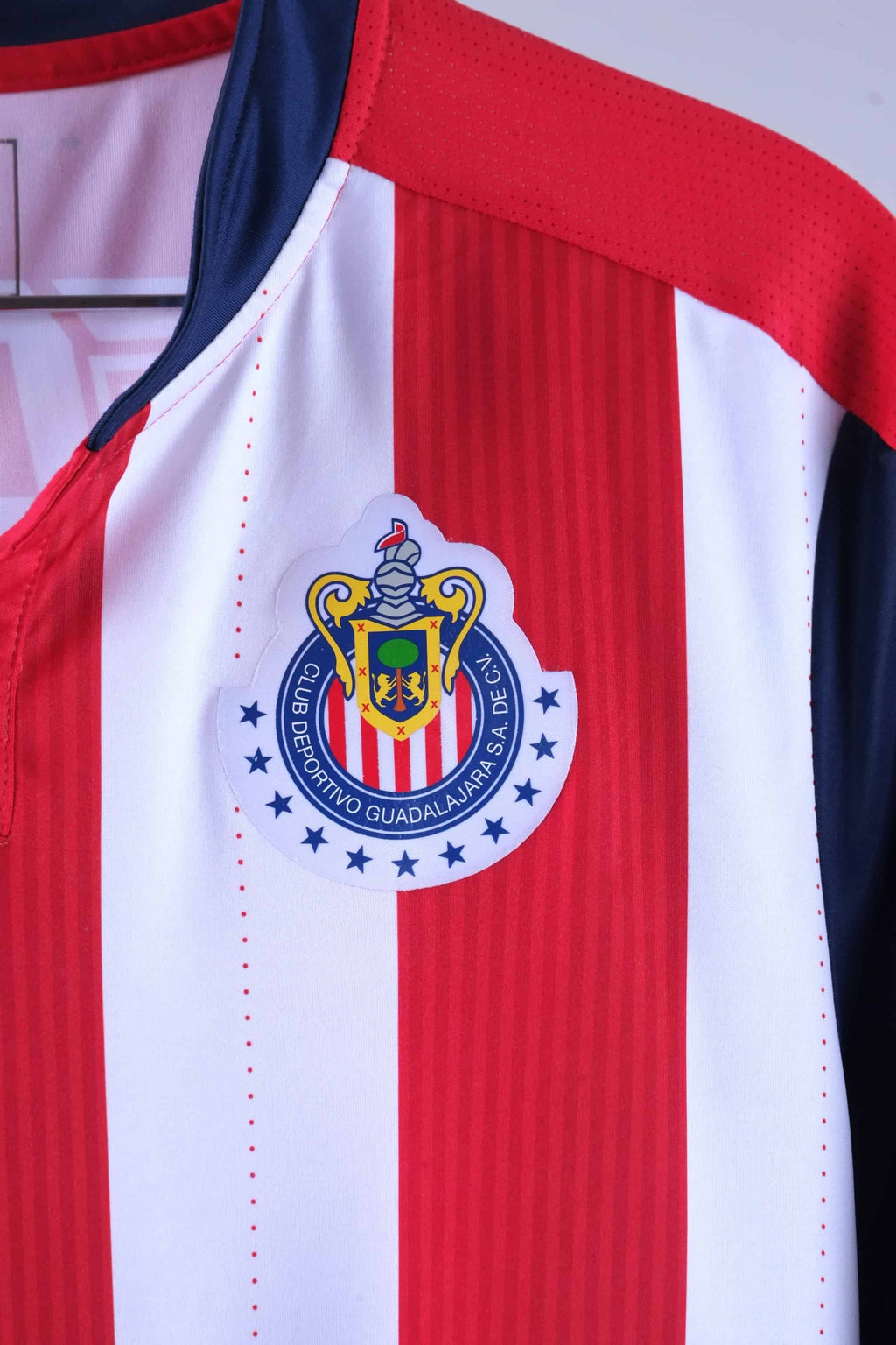 Close-up shot of the circular Chivas de Guadalajara club crest heat-pressed onto the red and white striped fabric, surrounded by blue stars.