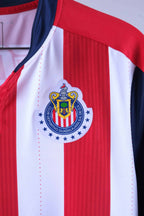 Close-up shot of the circular Chivas de Guadalajara club crest heat-pressed onto the red and white striped fabric, surrounded by blue stars.