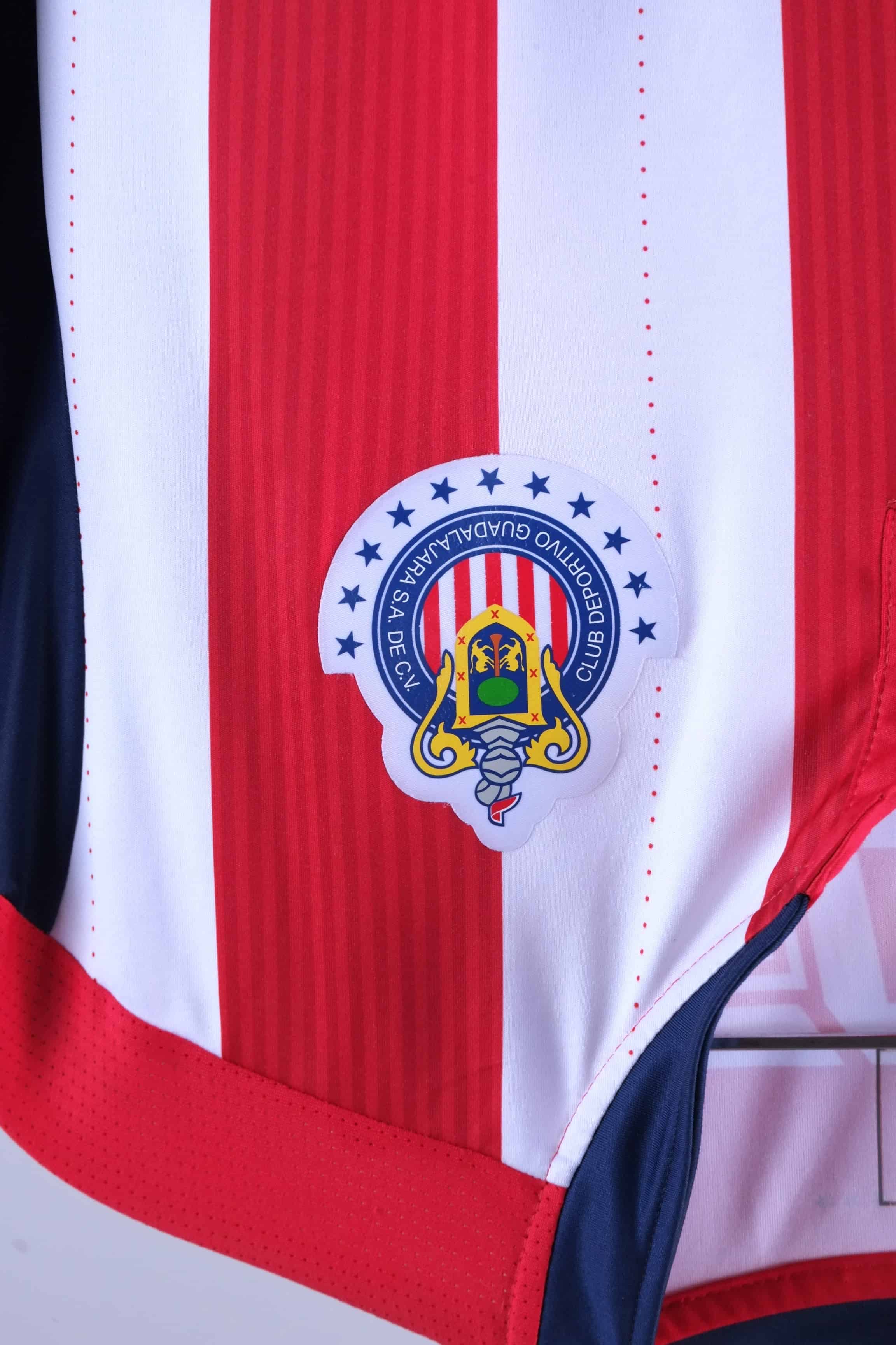 Close-up shot of the circular Chivas de Guadalajara club crest heat-pressed onto the red and white striped fabric, surrounded by blue stars.